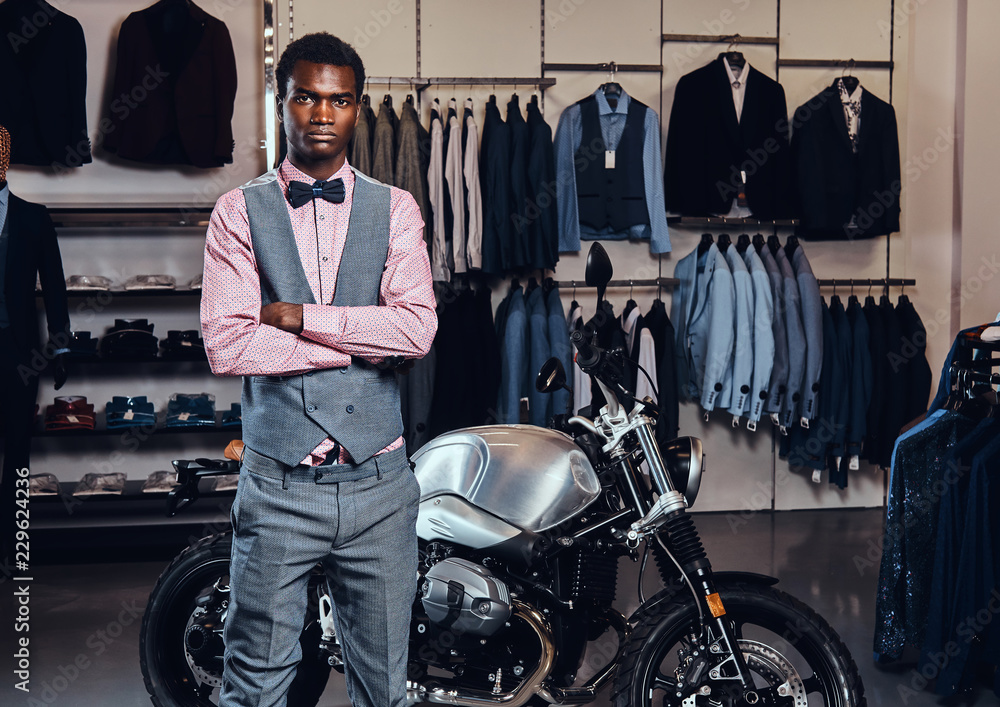 Elegantly dressed African American young man posing with crossed arms near retro sports motorbike at the men's clothing store.