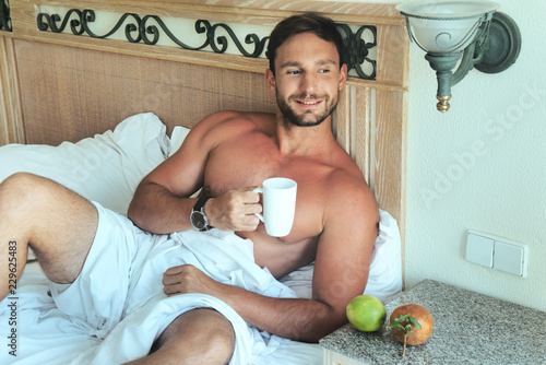 Attractive young bachelor man lying in bed with naked muscular torso and a sexy look with a smirk on his handsome face enjoing his morning. isolated on white background with big biceps and chest