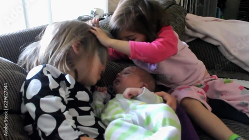 Two sisters fight over who gets to kiss newborn baby brother.  One is holding and kissing him, the other pushes her head back and leans in to kiss him herself.  Home video of kids. photo