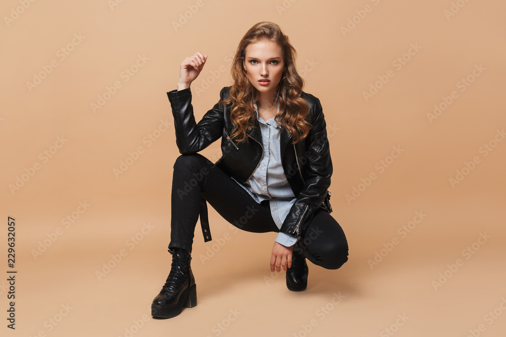 Beautiful serious girl with wavy hair in black leather jacket and shirt thoughtfully looking in camera over beige background isolated - obrazy, fototapety, plakaty 