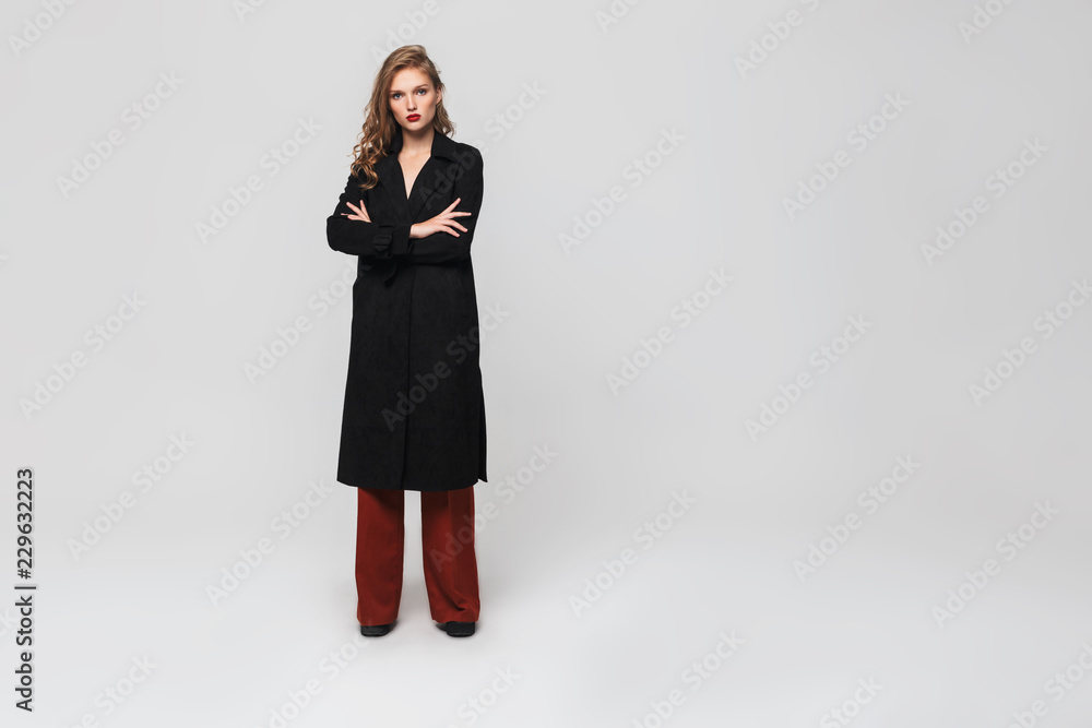 Young attractive pensive woman with wavy hair in black coat and red trousers holding hands folded while thoughtfully looking in camera over gray background Young attractive pensive woman with wavy hair in black coat and red trousers holding hands folded while thoughtfully looking in camera over gray background