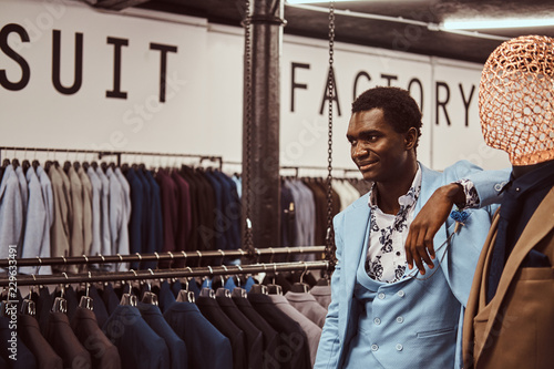 Elegantly dressed African-American man leaning on mannequin in a classic menswear store.