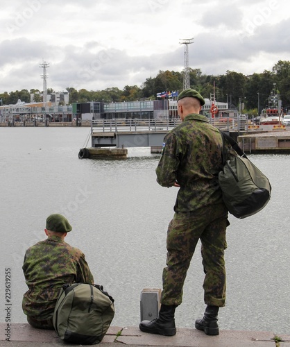 Two young conscripts in the Finnish army on temporary leave stand in uniform looking out to the harbor in Helsinki, Finland. Portrait format photo.