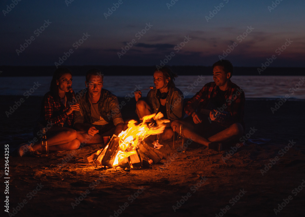 Camp on the beach. Group of young friends having picnic with bonfire. They eat sausages Camp on the beach. Group of young friends having picnic with bonfire. They eat sausages