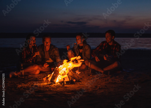 Camp on the beach. Group of young friends having picnic with bonfire. They eat sausages