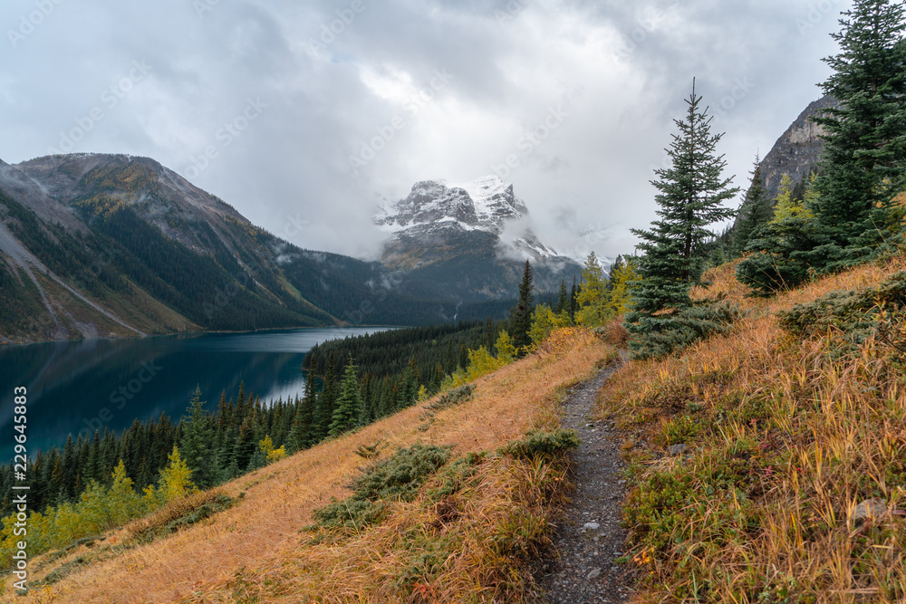 A trail follows the ridge above a blue alpine lake in the Rockies