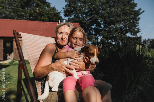 Smiling grandmother and granddaughter with dog sitting on deckchair in garden photo