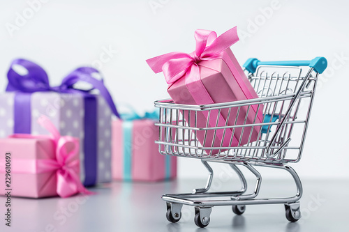 Shopping cart with gift box in pink packaging. Gifts in multi-colored packaging, decorated with ribbons and bows.