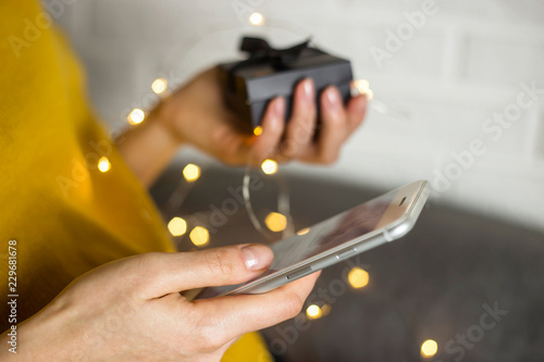 The girl is holding a smartphone with which she orders gifts from online stores from home. Cyber Monday and Black Friday concept. Gift box in hand with lights