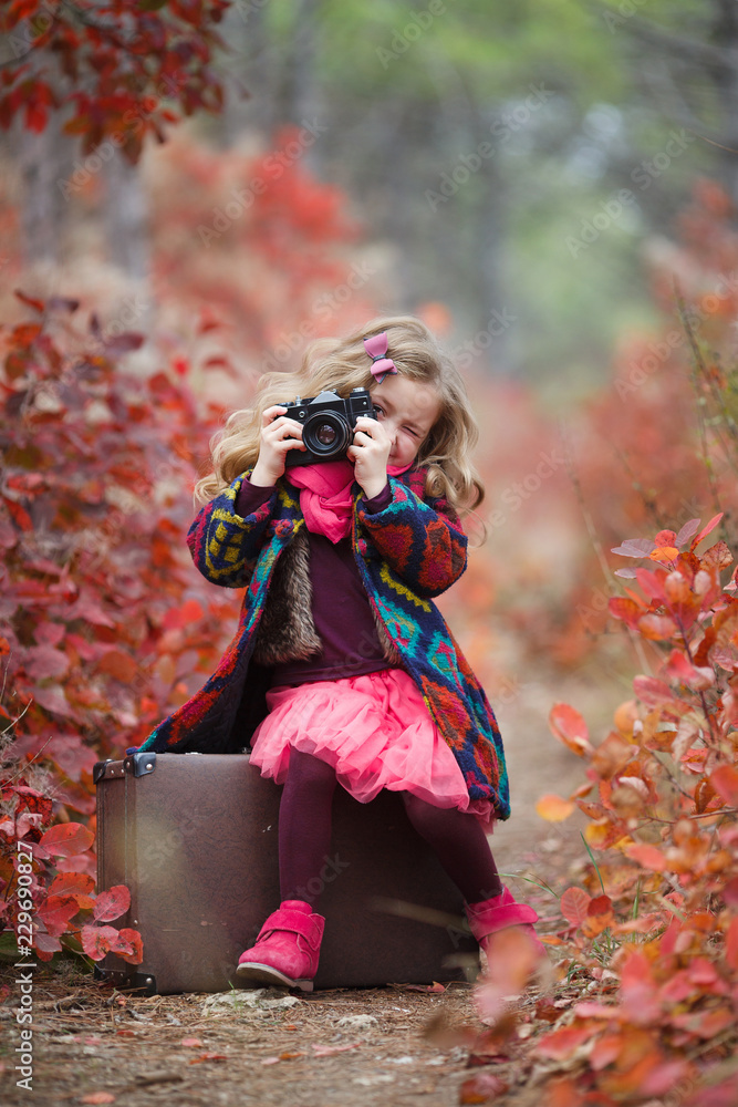 Little cute girl in an autumn jacket photographs the golden autumn,standing on the road in the woods with an old suitcase.Little kid is playing with camera and taking photo of autumn nature as master. Little cute girl in an autumn jacket photographs the golden autumn,standing on the road in the woods with an old suitcase.Little kid is playing with camera and taking photo of autumn nature as master.