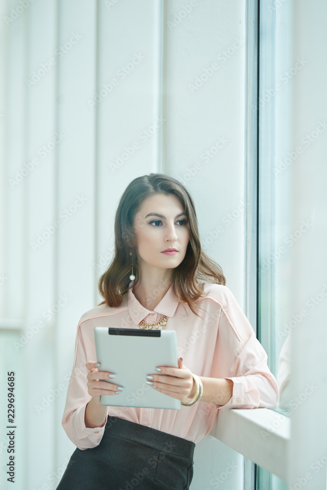 Pensive young female entrepreneur with tablet computer standing at office window Pensive young female entrepreneur with tablet computer standing at office window