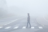 Young man alone without reflector elements crossing street on crosswalk in mist of early morning. Poor visibility. Foggy air. Side view.