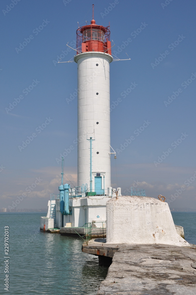 Lighthouse of Odessa port in Ukraine. Antique white beacon with seascape background. Red lantern lamp on the top of lighthouse building with clear blue sky background.