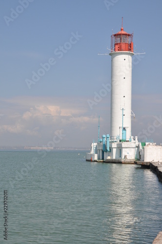 Lighthouse of Odessa port in Ukraine. Old white beacon with reflection in calm sea water surface and seascape background. Red lantern lamp on the top of lighthouse building. Black sea view