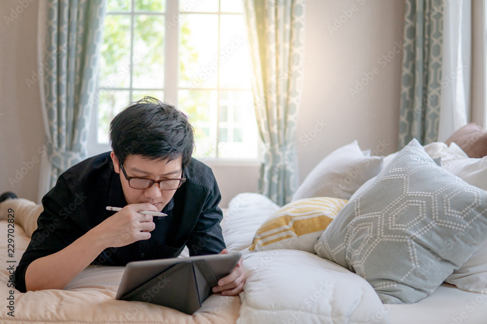 Young Asian business man using digital tablet and pen while lying on bed in cozy bedroom. Home living lifestyle with modern electronic gadget concept Young Asian business man using digital tablet and pen while lying on bed in cozy bedroom. Home living lifestyle with modern electronic gadget concept