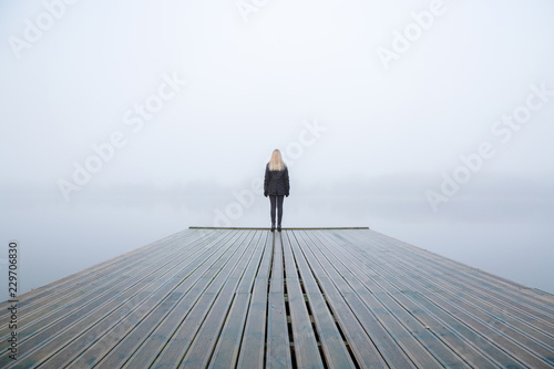 Young woman standing alone on edge of footbridge and staring at lake. Mist over water. Foggy air. Early chilly morning in autumn. Beautiful freedom moment and peaceful atmosphere in nature. Back view.