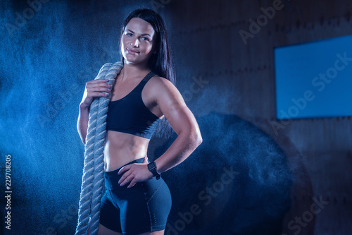 Strong beautiful woman with a battle rope on her shoulder in the gym.