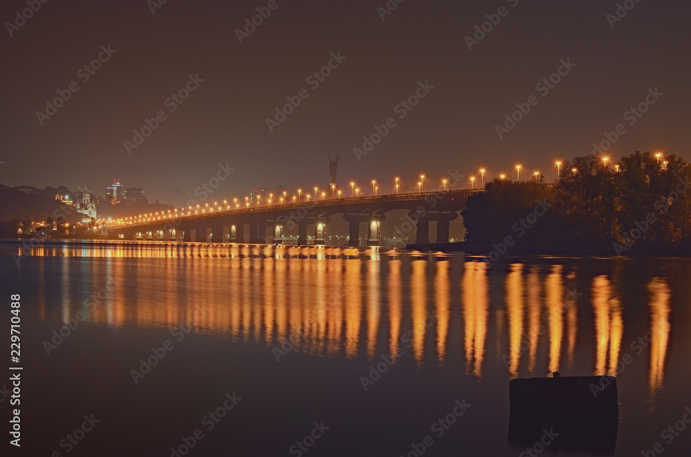 Autumn night cityscape with Paton bridge over Dnieper river. Motherland monument at the background. City lights reflected in the water. Kyiv, Ukraine