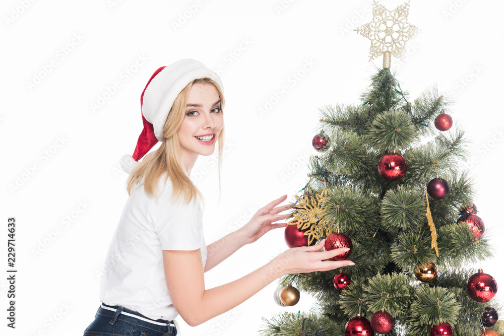 side view of smiling woman in santa claus hat decorating christmas tree isolated on white