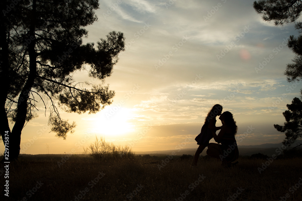 backlight of a mother kissing her daughter in a pine forest with yellow sky at sunset sunrise