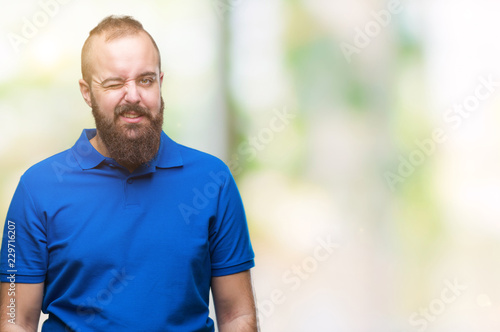Young caucasian hipster man wearing blue shirt over isolated background winking looking at the camera with sexy expression, cheerful and happy face. © Krakenimages.com