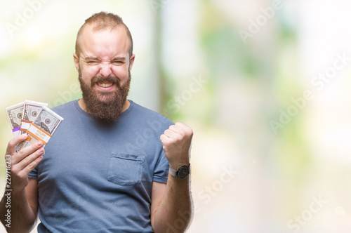 Young hipster man holding bunch of money over isolated background screaming proud and celebrating victory and success very excited, cheering emotion