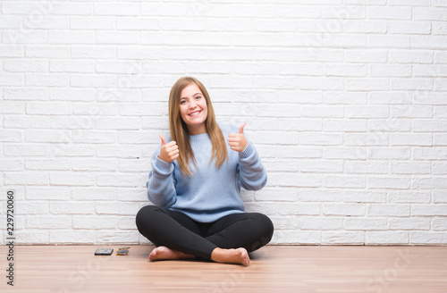 Young adult woman sitting on the floor in autumn over white brick wall success sign doing positive gesture with hand, thumbs up smiling and happy. Looking at the camera with cheerful expression