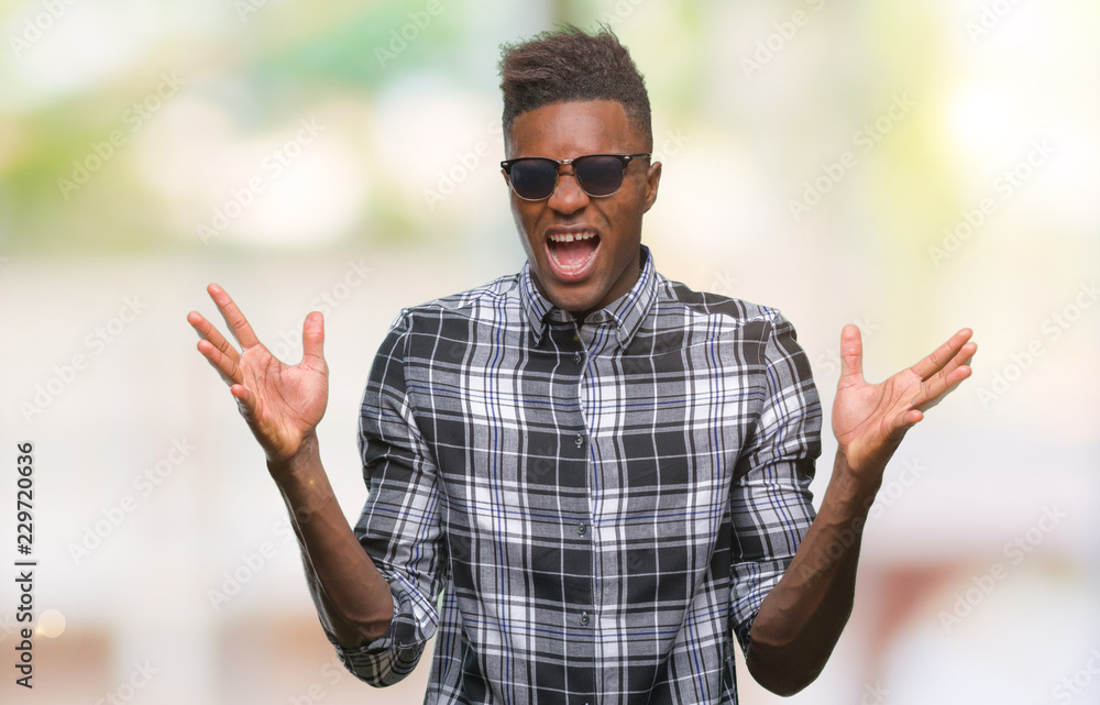 Naklejka premium Young african american man wearing sunglasses over isolated background celebrating crazy and amazed for success with arms raised and open eyes screaming excited. Winner concept