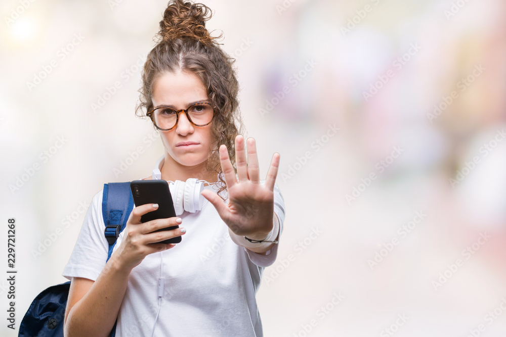 Young brunette student girl wearing backpack, headphones and smartphone over isolated background with open hand doing stop sign with serious and confident expression, defense gesture