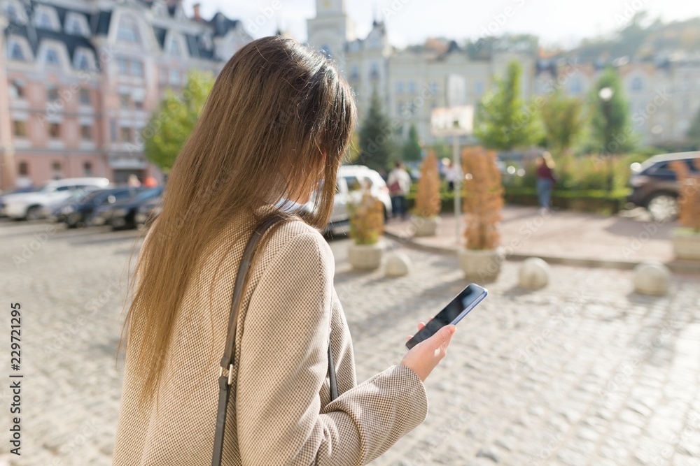 Young woman texting on the smart phone walking in the sunny city street, girl wearing warm coat, view from the back