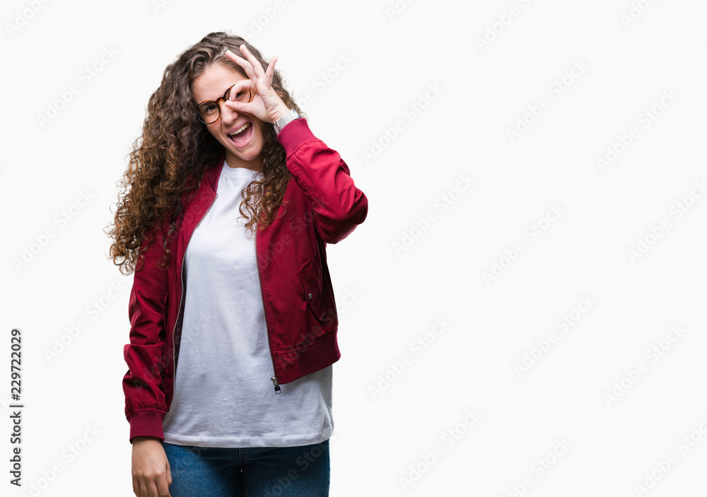 Beautiful brunette curly hair young girl wearing jacket and glasses over isolated background doing ok gesture with hand smiling, eye looking through fingers with happy face. Beautiful brunette curly hair young girl wearing jacket and glasses over isolated background doing ok gesture with hand smiling, eye looking through fingers with happy face.