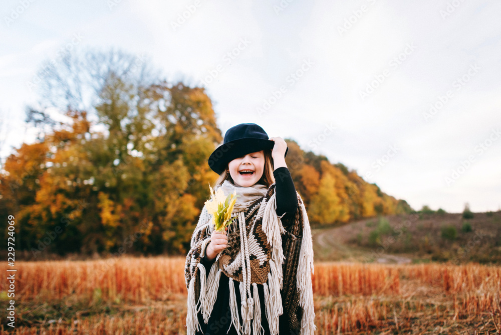 Very emotional autumn portrait of a cute little girl in a field near the forest. Atmospheric autumn background. Little cute girl in a poncho and a hat in a field with several leaves in her hand. 