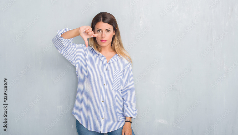 Young adult woman over grey grunge wall wearing fashion business outfit looking unhappy and angry showing rejection and negative with thumbs down gesture. Bad expression. Young adult woman over grey grunge wall wearing fashion business outfit looking unhappy and angry showing rejection and negative with thumbs down gesture. Bad expression.