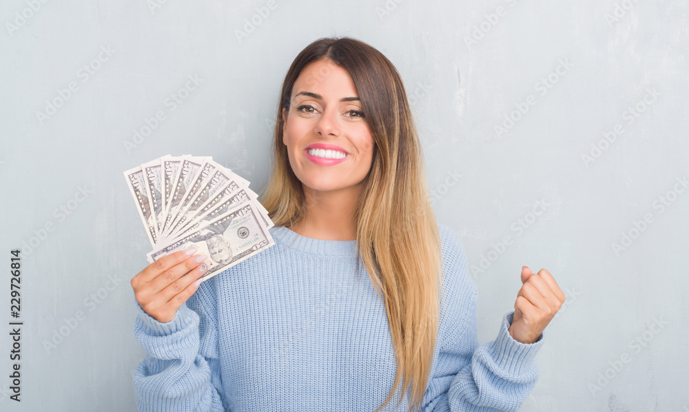 Young adult woman over grey grunge wall holding dollars screaming proud and celebrating victory and success very excited, cheering emotion Young adult woman over grey grunge wall holding dollars screaming proud and celebrating victory and success very excited, cheering emotion