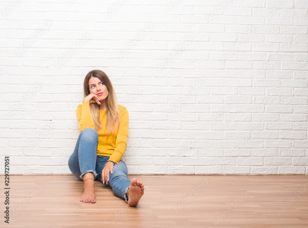 Young adult woman sitting on the floor over white brick wall with hand on chin thinking about question, pensive expression. Smiling with thoughtful face. Doubt concept. Young adult woman sitting on the floor over white brick wall with hand on chin thinking about question, pensive expression. Smiling with thoughtful face. Doubt concept.