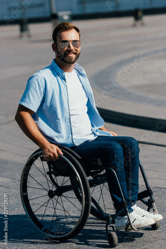 portrait of handsome man in sunglasses using wheelchair on street and looking at camera