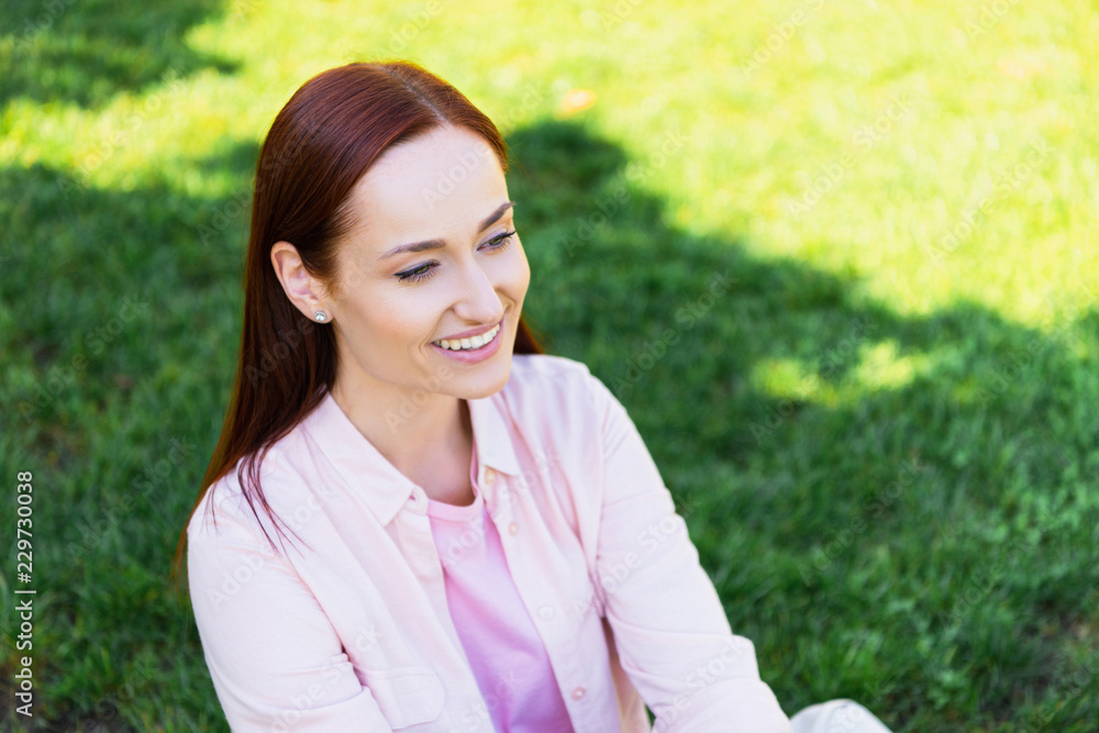 portrait of attractive smiling redhair woman in pink shirt looking away in park portrait of attractive smiling redhair woman in pink shirt looking away in park