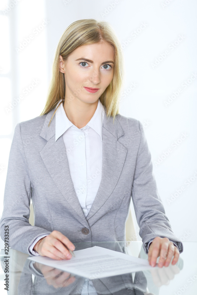 Blonde business woman examining contract while sitting at the glass desk in office background . Lawyer or secretary specialist at work Blonde business woman examining contract while sitting at the glass desk in office background . Lawyer or secretary specialist at work