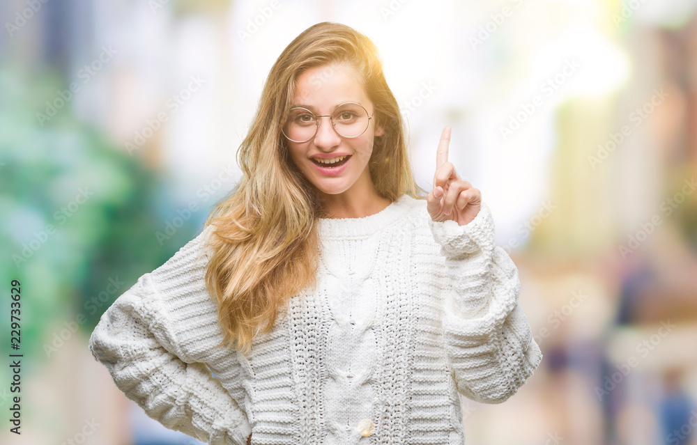 Young beautiful blonde woman wearing winter sweater and sunglasses over isolated background pointing finger up with successful idea. Exited and happy. Number one.