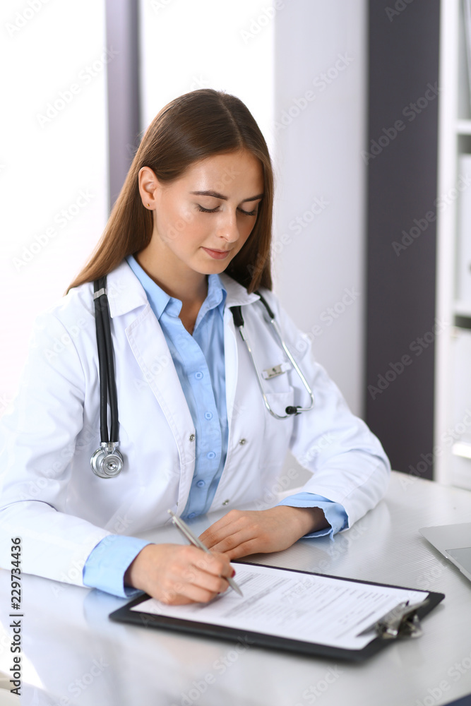 Doctor woman filling up medical form while sitting at the desk in hospital office. Physician at work. Medicine and health care concept Doctor woman filling up medical form while sitting at the desk in hospital office. Physician at work. Medicine and health care concept