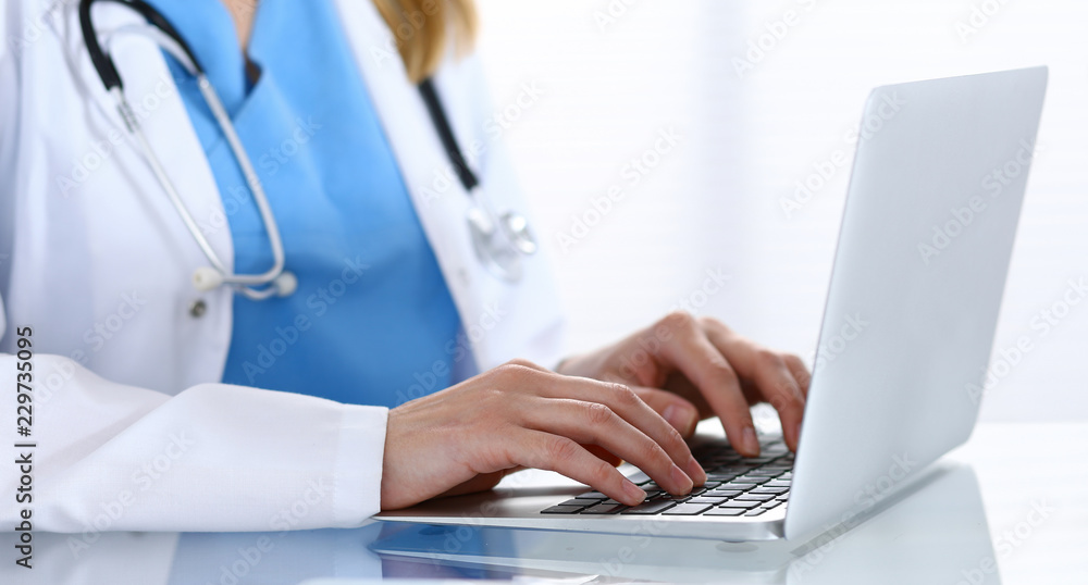Doctor typing on laptop computer while sitting at the glass desk in hospital office. Physician at work. Medicine and healthcare concept Doctor typing on laptop computer while sitting at the glass desk in hospital office. Physician at work. Medicine and healthcare concept