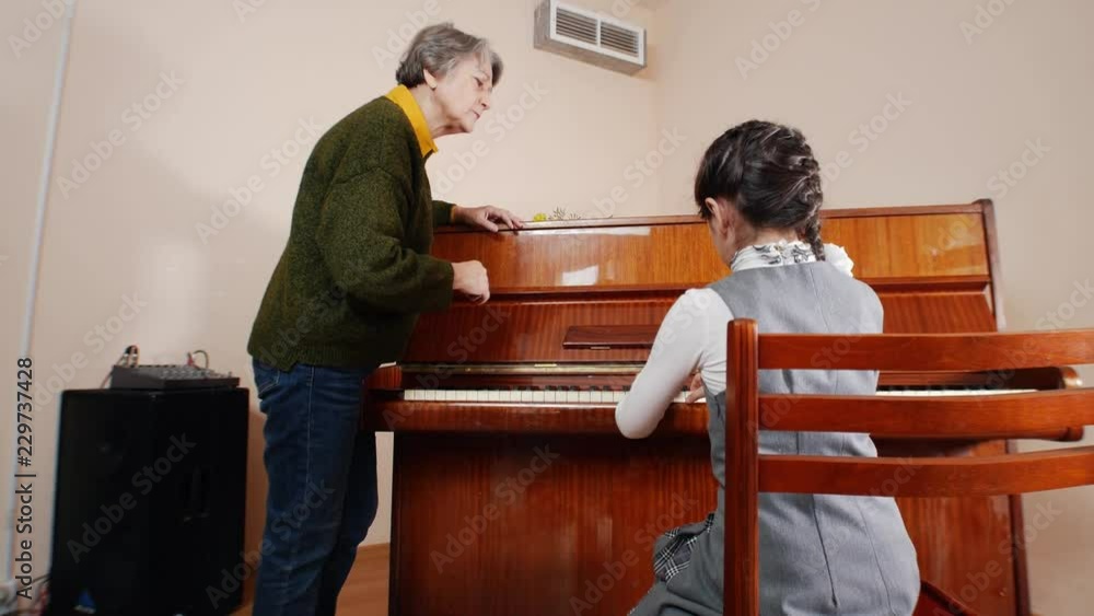 Music lesson. Girl playing piano, elder teacher stands near the piano and helps with playing on the piano. Slider view of playing from back of the girl Music lesson. Girl playing piano, elder teacher stands near the piano and helps with playing on the piano. Slider view of playing from back of the girl