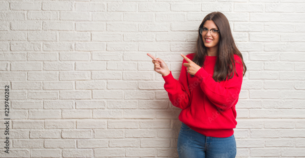 Young brunette woman standing over white brick wall smiling and looking at the camera pointing with two hands and fingers to the side. Young brunette woman standing over white brick wall smiling and looking at the camera pointing with two hands and fingers to the side.
