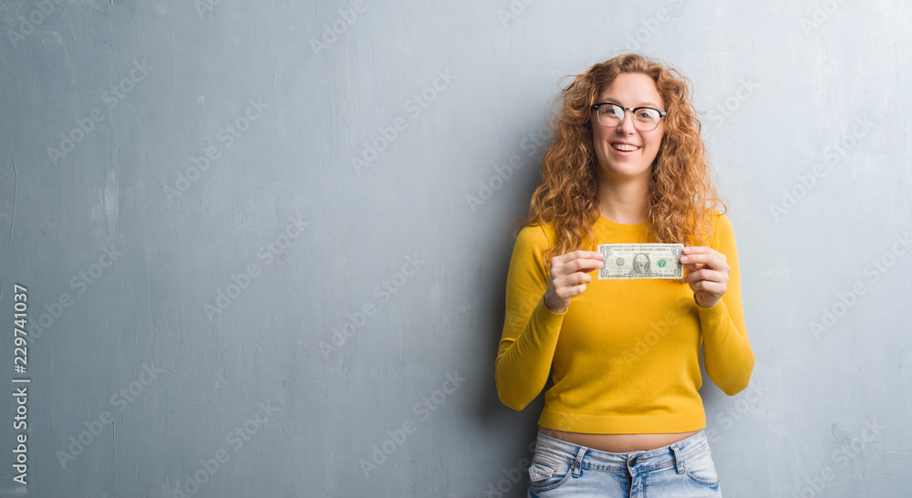 Young redhead woman over grey grunge wall holding a dollar with a happy face standing and smiling with a confident smile showing teeth Young redhead woman over grey grunge wall holding a dollar with a happy face standing and smiling with a confident smile showing teeth