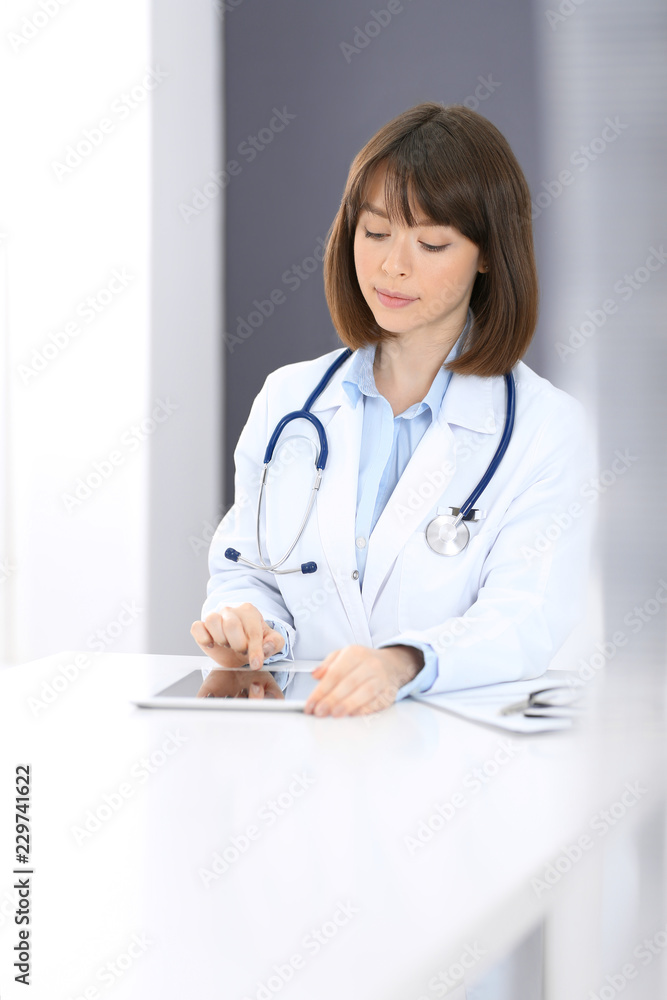 Female doctor typing on laptop computer while sitting at the white table in hospital office. Physician at work. Medicine and healthcare concept Female doctor typing on laptop computer while sitting at the white table in hospital office. Physician at work. Medicine and healthcare concept