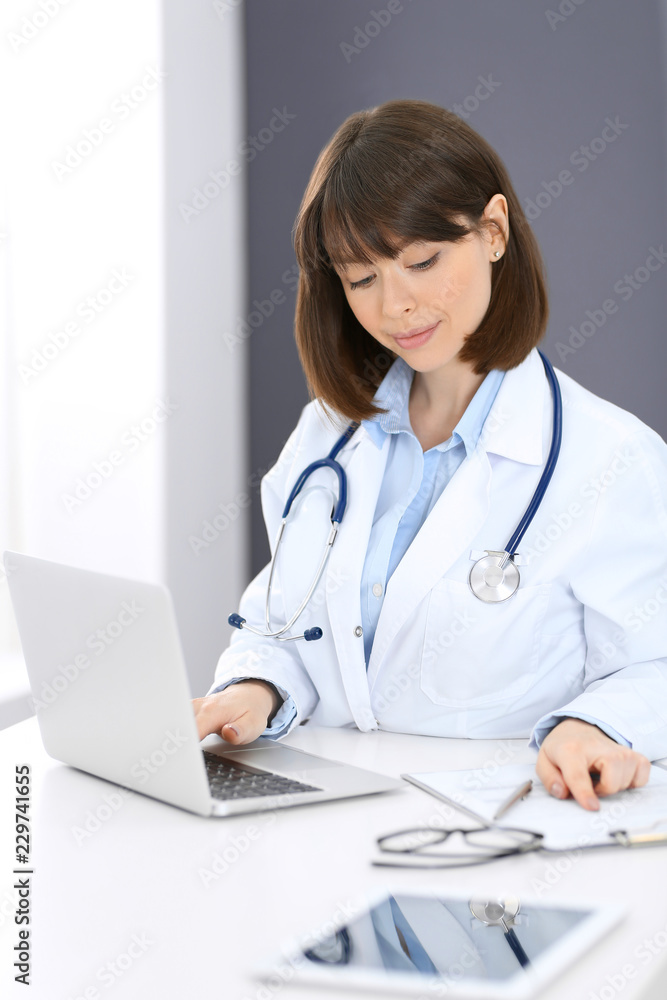 Doctor typing on laptop computer while sitting at the white table in hospital office. Physician at work. Medicine and healthcare concept Doctor typing on laptop computer while sitting at the white table in hospital office. Physician at work. Medicine and healthcare concept