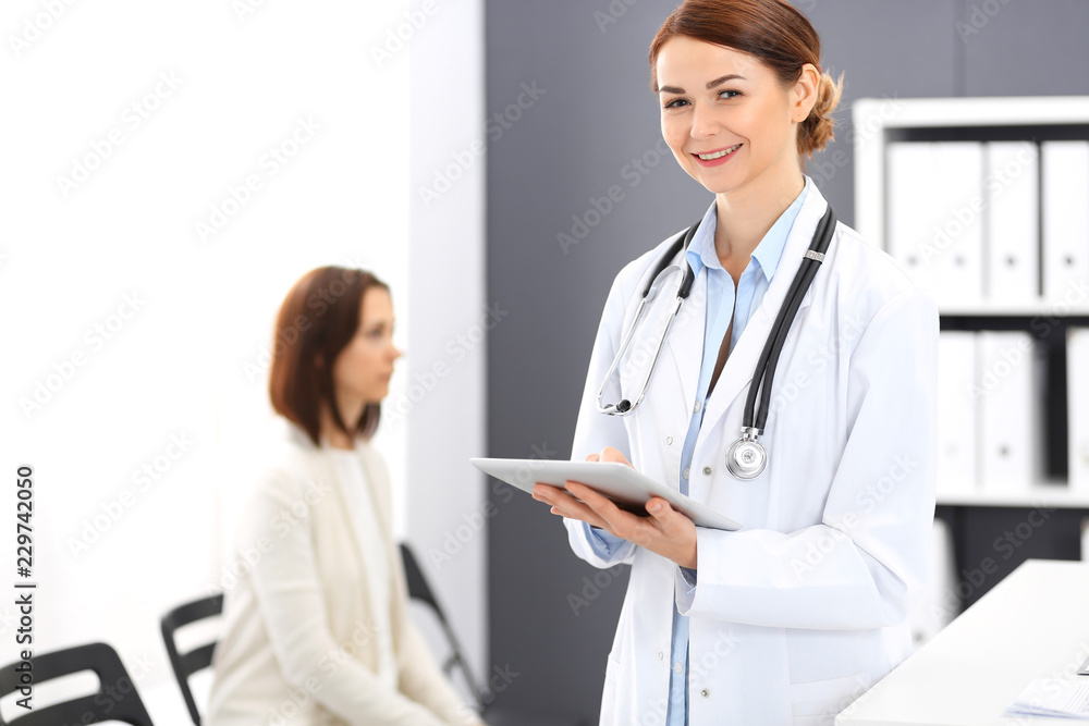 Happy doctor woman at work. Portrait of female physician using tablet computer while standing near reception desk at clinic or emergency hospital. Patient woman sitting at the background. Medicine Happy doctor woman at work. Portrait of female physician using tablet computer while standing near reception desk at clinic or emergency hospital. Patient woman sitting at the background. Medicine