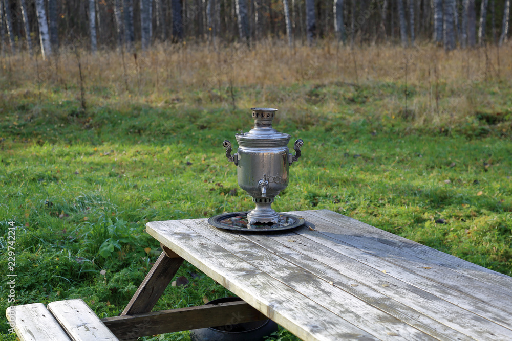 A device for boiling water and making tea. Traditional Russian samovar on a wooden table against the background of autumn nature
A device for boiling water and making tea. Traditional Russian samovar on a wooden table against the background of autumn nature