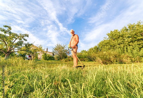 The gray-haired man mows the grass with a trimmer on a sunny day. photo