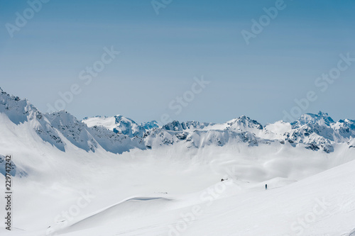The mountaineer stands on the edge of the glacier with a snow shovel in his hands and shows Shak's gesture against the blue sky and the Caucasus Mountains
